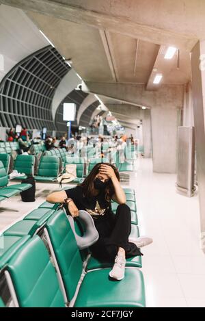 Von oben besorgte junge Frau mit gekreuzten Beinen, die eine Maske trug und den Kopf berührte, die auf einem Stuhl im Flughafen saß Stockfoto