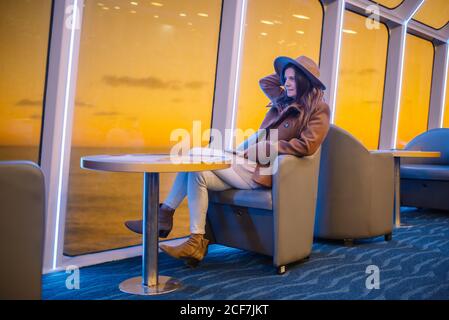 Entspannt stilvolle junge Frau im trendigen Outfit auf Stuhl sitzend An Bord eines Luxuskreuzfahrtschiffes und genießen Sie die Aussicht vom Fenster mit Ruhiges Meerwasser und Sonnenuntergang Himmel Stockfoto