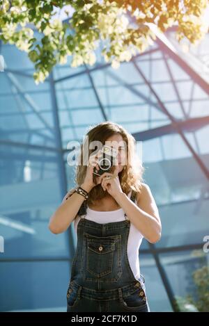 Junge, enthusiastische Frau, die auf dem Hintergrund der Glasarchitektur Momente mit der Kamera fotografiert Stockfoto