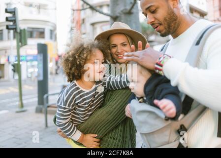 Positive stilvolle junge multirassische Eltern und nette kleine Tochter streicheln Kopf des Jungen vom Vater getragen, während sie zusammen stehen An der City Street Stockfoto