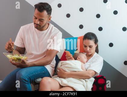 Schwarz haarige Mutter mit schläfrigen Baby ob Brust mit Handy Telefon in der Hand sitzend auf Stuhl mit Vater in der Freizeit Tragen Sie essenden Salat Stockfoto