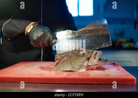 Unkenntlich garen in schwarzer Uniform und Handschuhe mit Metallspalter Beim Schneiden Kopf von großen frischen Fisch auf rot gehackt Tafel am Tisch gegen verschwommenes Interieur der modernen Küche Stockfoto