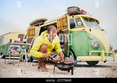 Aufmerksamer älterer Mann in legerer Kleidung, der auf dem Campingkocher kocht Neben serviert auf der Straße Tisch und geparkten Wohnmotorhaus Am Sommertag Stockfoto