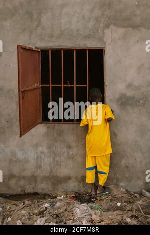 Gambia, Afrika - 3. August 2019: Rückansicht eines afrikanischen Jungen in hellgelber Kleidung, der in ein dunkles Fenster guckt, mit einem Gitter eines alten Gebäudes, das auf Müllland steht Stockfoto