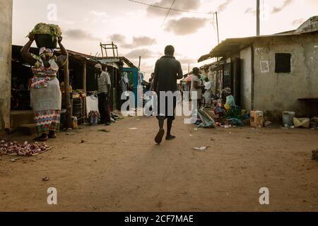 Gambia, Afrika - 3. August 2019: Rückansicht des gesichtslosen armen schwarzen Mannes, der auf schmutziger überfüllter Straße zwischen den Theken und Häusern des afrikanischen Dorfes läuft Stockfoto