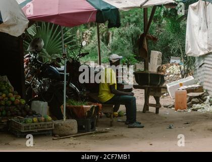 Gambia, Afrika - 5. August 2019: Seitenansicht eines ruhigen afrikanischen Verkäufers, der auf einer Holzbank in der Nähe von Motorrädern unter einem kleinen Zelt mit Früchten sitzt, während er auf die Kunden wartet, die wegschauen Stockfoto