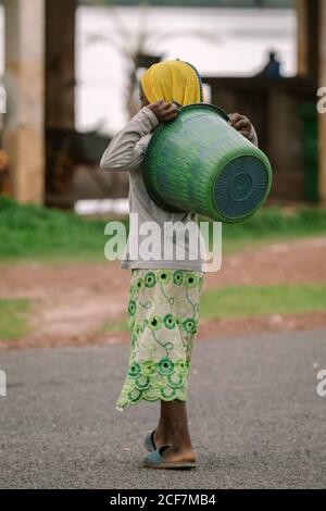 Gambia, Afrika - 7. August 2019: Rückansicht eines kleinen armen Kindes in einfacher Kleidung, das während des Spaziergangs auf der Straße grünes Becken auf der Rückseite hält Stockfoto