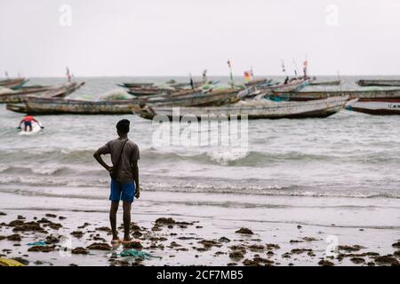 Gambia, Afrika - 6. August 2019: Rückansicht eines schwarzen Mannes, der am schmutzigen Strand steht und die Fischerboote tagsüber an der Küste ansieht Stockfoto