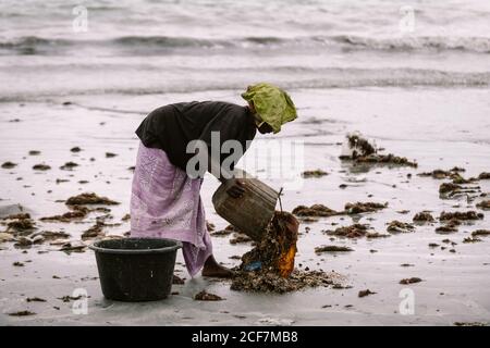 Gambia, Afrika - 6. August 2019: Seitenansicht einer schwarzen Frau, die schmutziges Wasser mit Kleidung aus dem Eimer auf den Boden während des Waschens an Land gießt Stockfoto