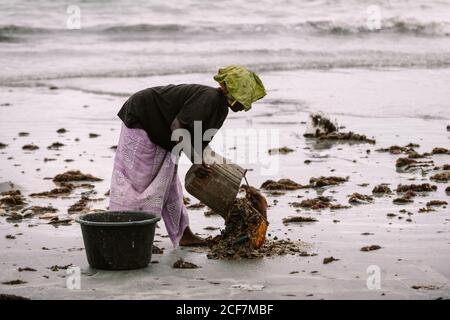 Gambia, Afrika - 6. August 2019: Seitenansicht einer schwarzen Frau, die schmutziges Wasser mit Kleidung aus dem Eimer auf den Boden während des Waschens an Land gießt Stockfoto