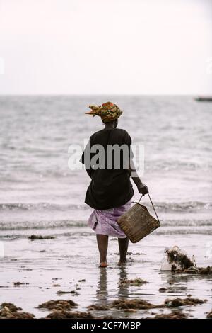 Gambia, Afrika - 6. August 2019: Seitenansicht einer schwarzen Frau, die schmutziges Wasser mit Kleidung aus dem Eimer auf den Boden während des Waschens an Land gießt Stockfoto