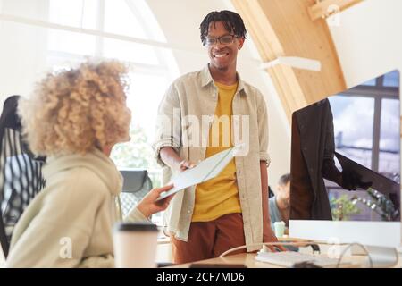 Afrikanischer Geschäftsmann in lässiger Kleidung lächelnd, während sie die Mappe Mit Dokumenten an seinen Kollegen, der am Tisch arbeitet Stockfoto