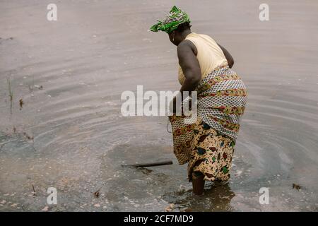 Gambia, Afrika - 7. August 2019: Rückansicht einer nicht erkennbaren schwarzen Frau in traditioneller Kleidung, die unbekannte Naturgegenstände im Rocksaum im flachen Wasser sucht und sammelt Stockfoto