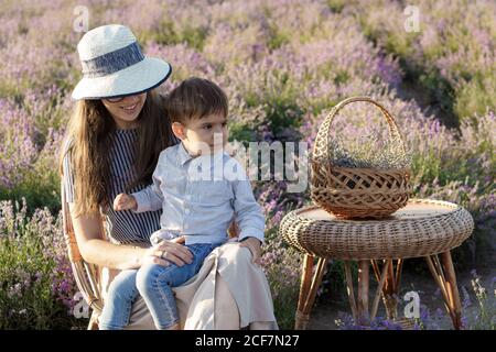 Große Familie, Vaterschaft, Kindheit, Mutterschaft, provence Stil Konzept - junge Mutter sitzt auf Korbstuhl mit kleinen Jungen Sohn mit Korb und Tisch Stockfoto