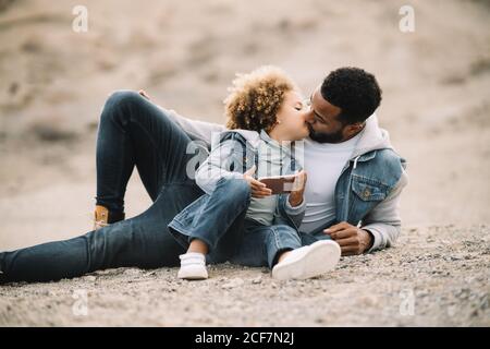 Afroamerikanischer bärtiger Casual-Mann, der auf sandigem Boden liegt und sich lehnt Am Ellenbogen und am Knie biegbar, mit Blick auf die Locken Ethnische Kleinkind in Denim-Kleidung sitzt neben mit Handy Stockfoto