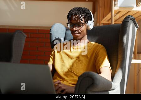 Afrikanischer Mann in kabellosen Kopfhörern, der auf einem Sessel sitzt und arbeitet Online auf Laptop-Computer Stockfoto