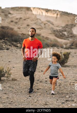 Afroamerikanischer bärtiger aktiver Vater im roten T-Shirt Laufen Mit fröhlichem lockigen Kind auf Wüstenlandschaft in Backlit Stockfoto