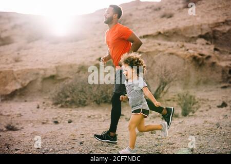 Afroamerikanischer bärtiger aktiver Vater im roten T-Shirt Laufen Mit fröhlichem lockigen Kind auf Wüstenlandschaft in Backlit Stockfoto