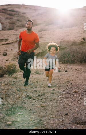 Afroamerikanischer bärtiger aktiver Vater im roten T-Shirt Laufen Mit fröhlichem lockigen Kind auf Wüstenlandschaft in Backlit Stockfoto