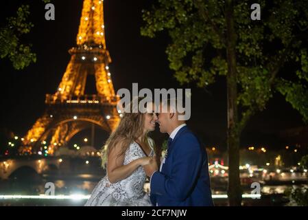 Joyful groom in blue suit and bride in white wedding dress holding hands while smiling and kissing on evening with Eiffel Tower on background at Paris Stockfoto