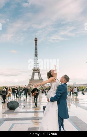 Bräutigam im blauen Anzug Heben fröhliche Braut mit offenen Armen In weiß Hochzeitskleid mit Eiffelturm auf dem Hintergrund bei Paris Stockfoto