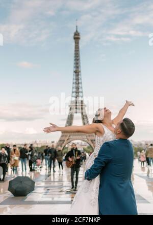 Bräutigam im blauen Anzug Heben fröhliche Braut mit offenen Armen In weiß Hochzeitskleid mit Eiffelturm auf dem Hintergrund bei Paris Stockfoto