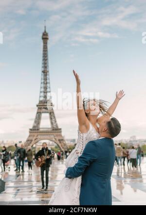 Bräutigam im blauen Anzug Heben fröhliche Braut mit offenen Armen In weiß Hochzeitskleid mit Eiffelturm auf dem Hintergrund bei Paris Stockfoto