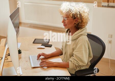 Reife Frau mit lockigen Haaren sitzt an ihrem Arbeitsplatz und Tippen Sie auf Computer-Tastatur und Blick auf Computer-Monitor sie Arbeiten mit Software Stockfoto