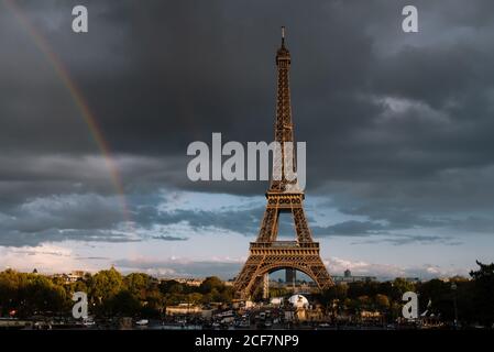 Mächtiger Eiffelturm zwischen grünen Pflanzen und Gebäuden mit bewölktem Himmel Himmel und Regenbogen auf dem Hintergrund in Paris Stockfoto