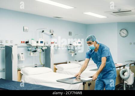 Medic in blau Uniform und Schutzmaske Einstellung Tablett auf Trolley im Krankenhauszimmer durch leere Betten Stockfoto