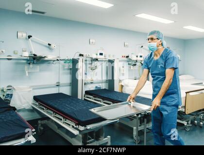 Medic in blau Uniform und Schutzmaske Einstellung Tablett auf Trolley im Krankenhauszimmer durch leere Betten Stockfoto