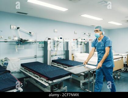 Medic in blau Uniform und Schutzmaske Einstellung Tablett auf Trolley im Krankenhauszimmer durch leere Betten Stockfoto