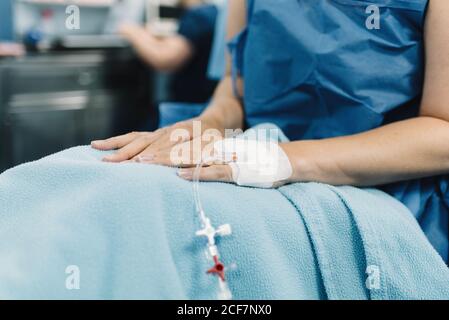 Crop weibliche Patientin sitzen mit bedeckten Beinen und intravenöser Flüssigkeit Nadel in der Hand vor der Operation im Operationssaal Stockfoto