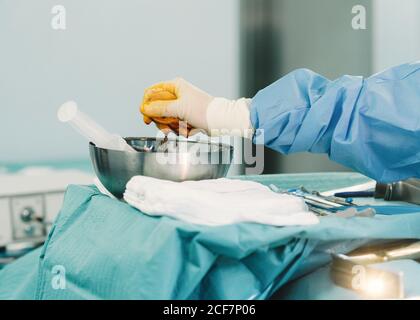Crop Hand von Sanitäter in blauem Kleid und weißen Handschuh Tampon mit Jod in der Schale während der Operation zusammendrücken Stockfoto