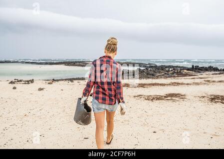 Positive Hipster Frau in legerer Kleidung und Handschuhen sammeln Müll in der Tasche, während hockt am einsamen Strand Stockfoto