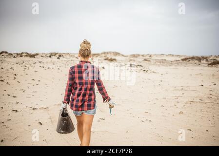 Positive Hipster Frau in legerer Kleidung und Handschuhen sammeln Müll in der Tasche, während hockt am einsamen Strand Stockfoto