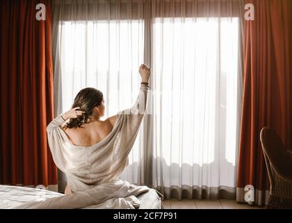 Rückansicht des erwachsenen Weibchens mit erhobenem Arm, der wegschaut Und Haare berühren, während man auf dem Bett gegen das geschlossene Fenster sitzt Mit roten Vorhängen und weißem Tüll am sonnigen Morgen Stockfoto