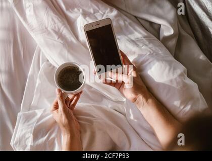 Von oben Crop unkenntlich Erwachsene weibliche Interaktion mit Smartphone während Sitzen im Bett und halten während Runde Tasse Kaffee Am sonnigen Morgen Stockfoto