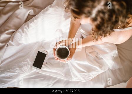 Von oben Crop unkenntlich Erwachsene weibliche Interaktion mit Smartphone während Sitzen im Bett und halten während Runde Tasse Kaffee Am sonnigen Morgen Stockfoto