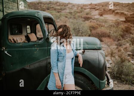 Junge Frau in Shorts und Jeanshemd, die neben einem grünen Lastwagen mit Holzkörper steht und auf den Hintergrund der Natur blickt Stockfoto