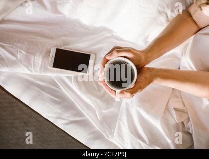 Von oben Crop unkenntlich Erwachsene weibliche Interaktion mit Smartphone während Sitzen im Bett und halten während Runde Tasse Kaffee Am sonnigen Morgen Stockfoto