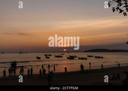 Silhouetten von nicht erkennbaren Menschen, die am Sandstrand und auf Booten spazieren Auf welligen Meer bei schönem Sonnenuntergang in Thailand Stockfoto