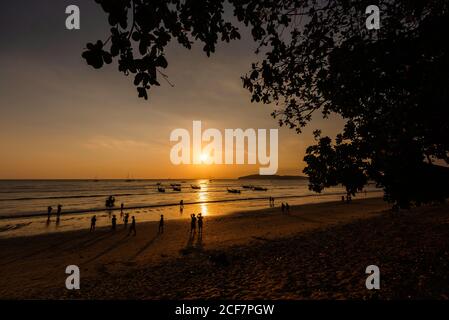 Silhouetten von nicht erkennbaren Menschen, die am Sandstrand und auf Booten spazieren Auf welligen Meer bei schönem Sonnenuntergang in Thailand Stockfoto