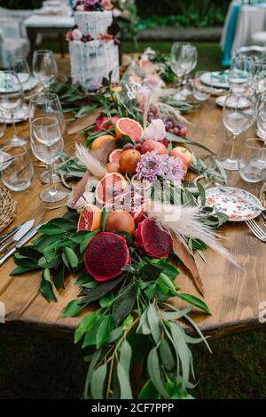 Schöne Girlande aus reifen geschnittenen tropischen Früchten und grün Zweige mit Blumen und dekorativen Pflanzen auf runden Holz Hochzeitstisch mit Tellern und Gläsern Stockfoto