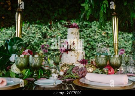 Weiße und goldene Stufenkuchen mit Blumen auf Hochzeit platziert Tisch mit Teller und Gläser mit Früchten und Blumen verziert Gegen grüne Bäume Stockfoto