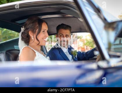 Glückliche Braut im Hochzeitskleid und Bräutigam in edlem Anzug Sitzen im Oldtimer und einander anschauen Stockfoto