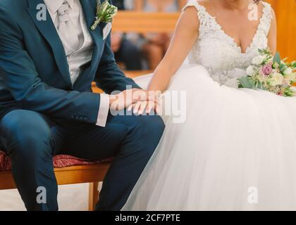 Unkenntlich Bräutigam im eleganten Anzug mit boutonniere und Braut in Weißes Hochzeitskleid sitzt auf der Bank und hält Hände während Zeremonie in der Kirche Stockfoto
