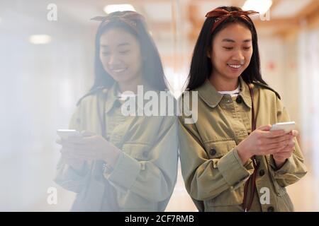 Asiatische junge Frau in lässiger Kleidung lächelnd beim Tippen ein Nachricht auf ihrem Handy Stockfoto
