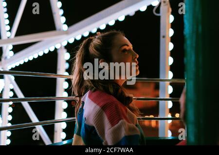Seitenansicht von verträumten Millennial weiblich im gestreiften Shirt genießen Blick vom Riesenrad während der Sommernacht im Vergnügen parken Stockfoto