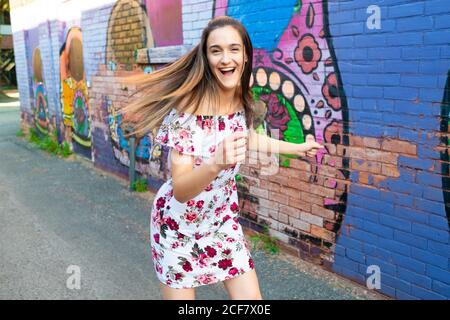 Verspielte Frau im Kleid, die Spaß hat, während sie mit Graffiti an der städtischen Ziegelmauer steht Stockfoto
